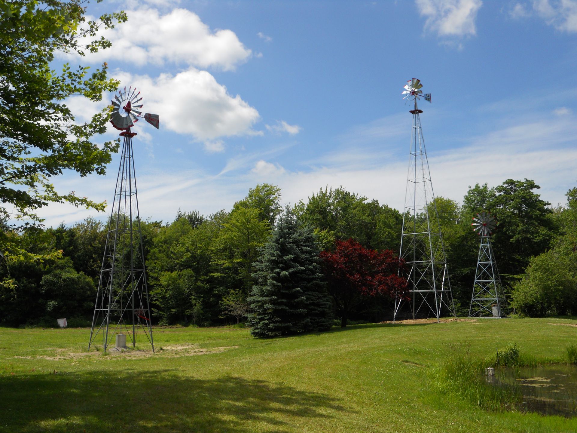 Three metal windmills stand in a grassy field, surrounded by trees under a blue sky with clouds.