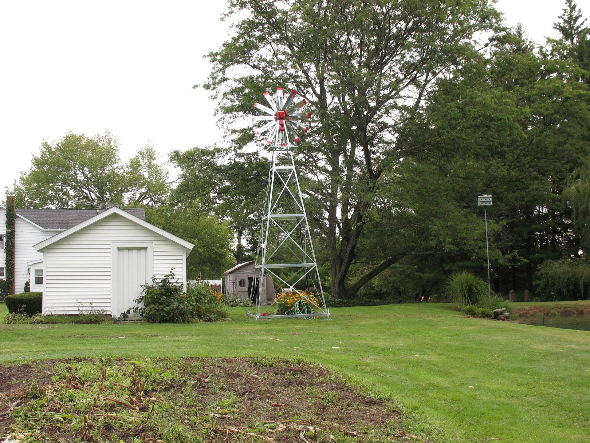 Windmill in a grassy yard, near a small white shed and trees, under a cloudy sky.