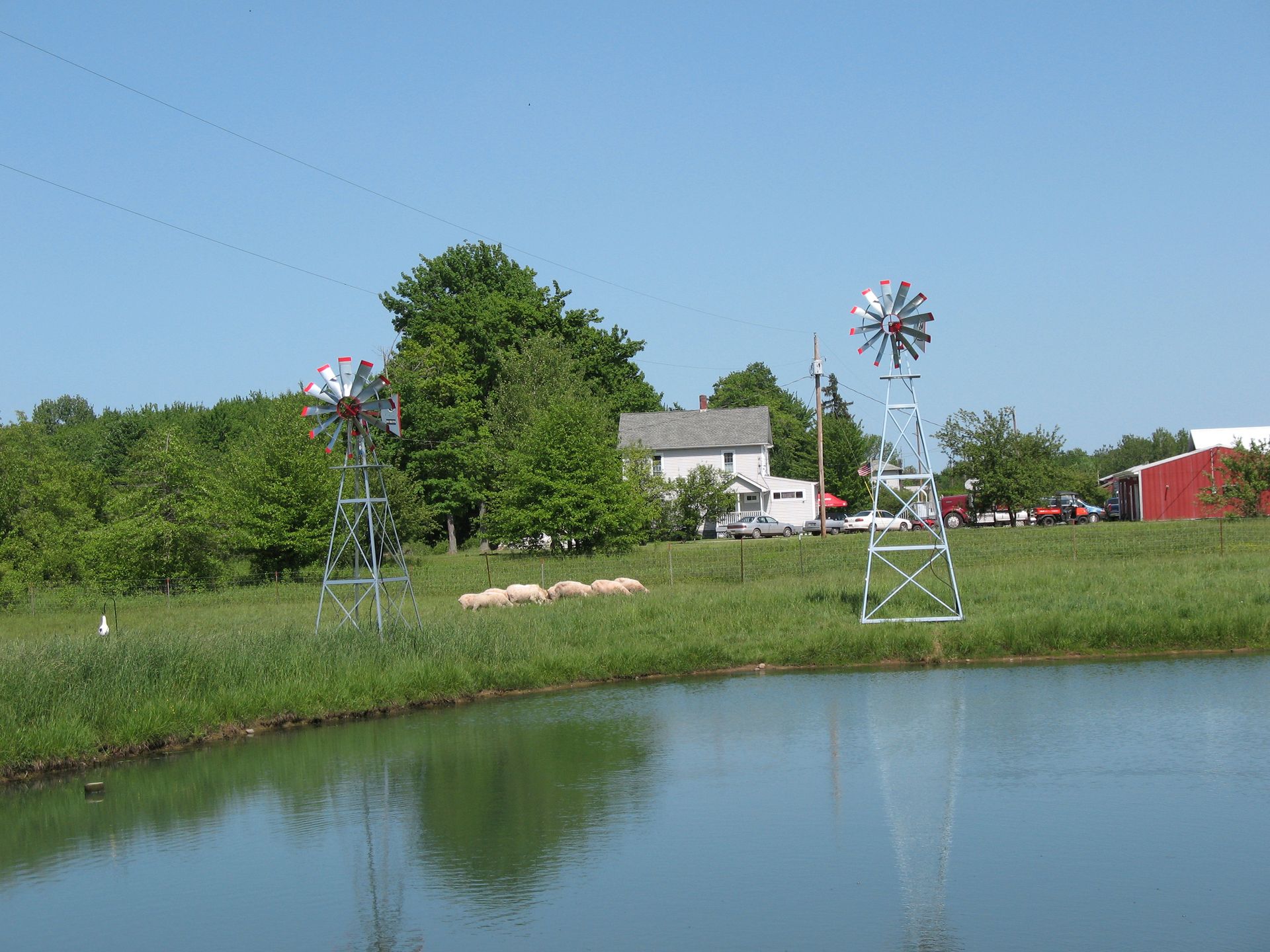 Two windmills stand on a grassy bank reflecting in a pond, with a house and trees in the background.