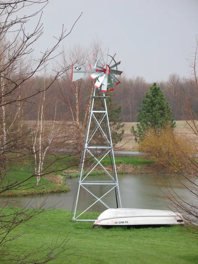 Windmill standing near a pond with a boat on the ground, trees in the background. Overcast day.