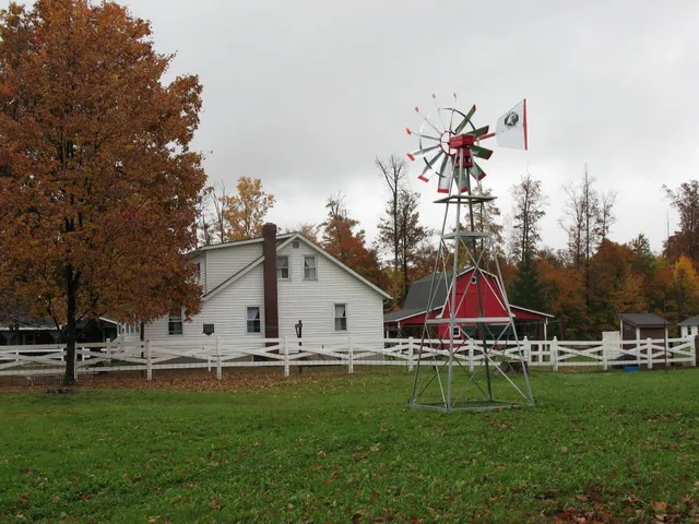 Farmhouse with red-trimmed windmill, white fence, and fall foliage under a cloudy sky.