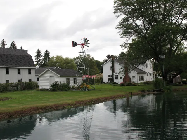 Waterfront rural scene: white houses, windmill, green grass, overcast sky, reflective water.