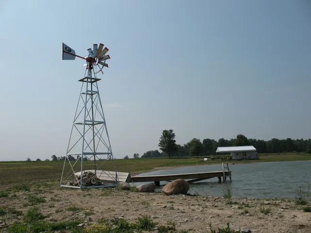 Windmill on a metal tower next to a wooden dock and a small white building by a lake.