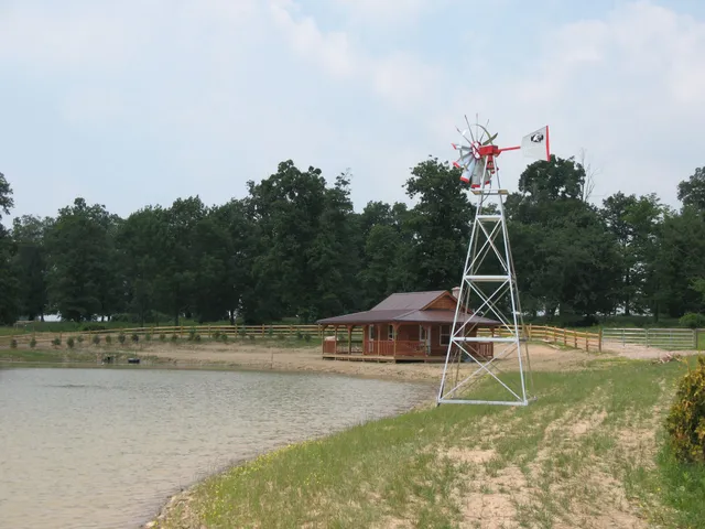 Windmill next to a small wooden building and pond, in front of a treeline under a cloudy sky.