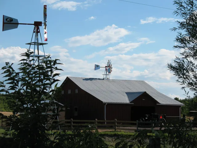 Barn with a red exterior and a silver roof, two windmills, and a cloudy blue sky.