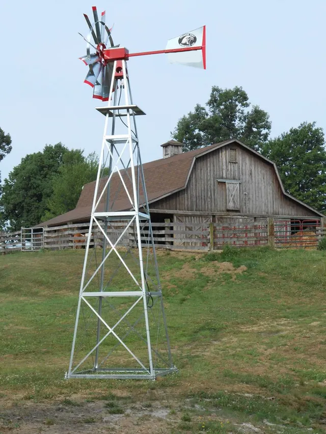 Windmill on a metal tower, with a red top, and a barn in the background on a grassy hill.