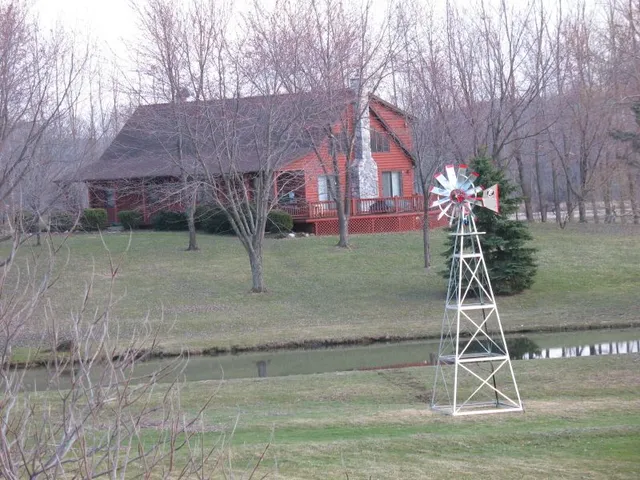 Red cabin with a windmill on a grassy lawn, near a water feature, trees in the background.