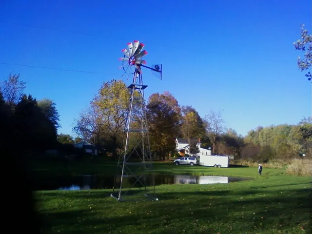Windmill on a tall metal tower near a pond and trees under a clear blue sky.