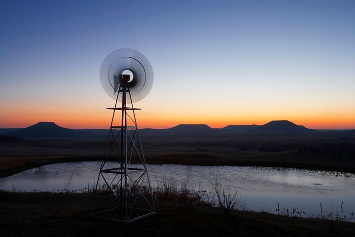Windmill silhouetted against an orange and blue sunset over a pond and hills.