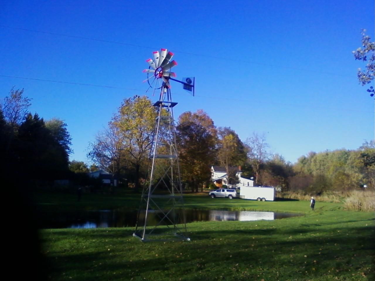 Windmill stands over a small pond on a sunny day with trees and a white vehicle in the background.