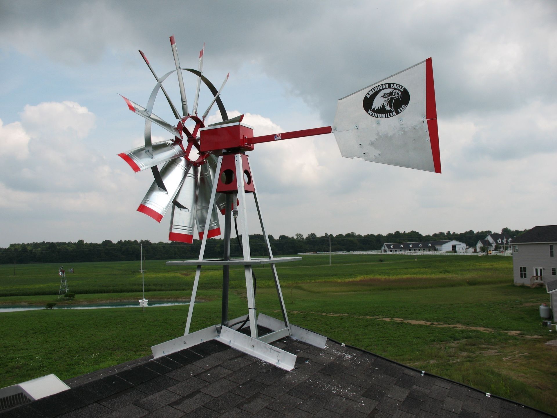 Windmill on a rooftop with red and silver blades and a white and red tail, set against a green field and sky.