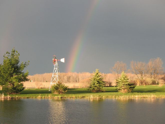 Rainbow arcs over a lake and windmill, fields, and trees, against a dark sky.