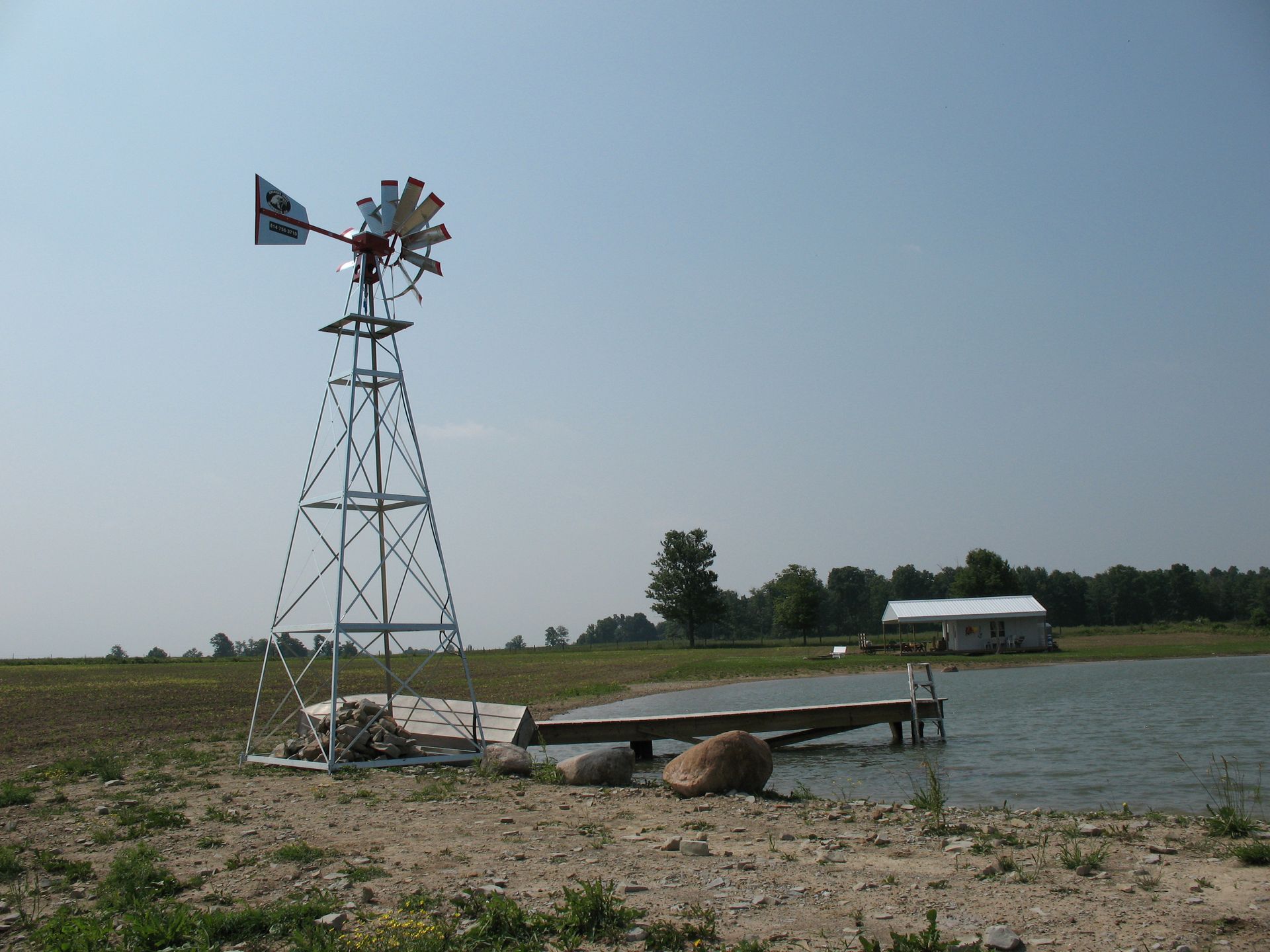 Windmill on a lake shore with a wooden dock and small building in the background.
