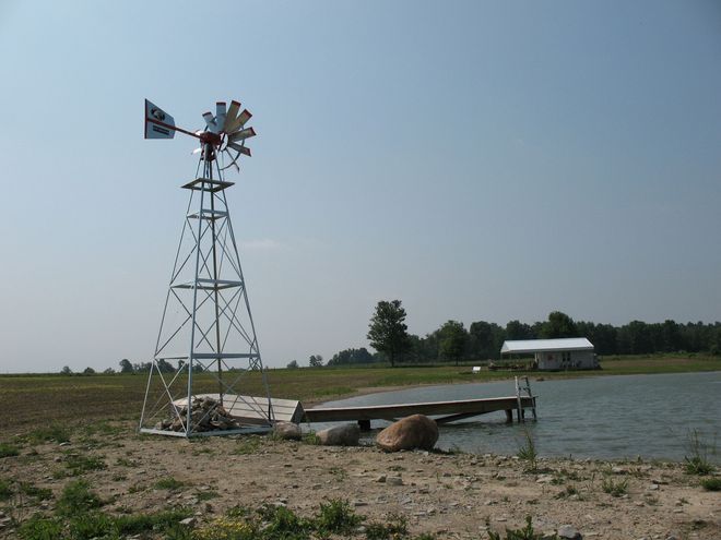 Windmill on a lake shore with a wooden dock and small building in the background.