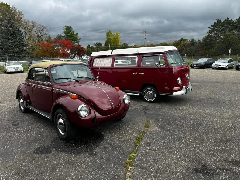 Burgundy Volkswagen Beetle convertible and van parked in a lot under cloudy skies.