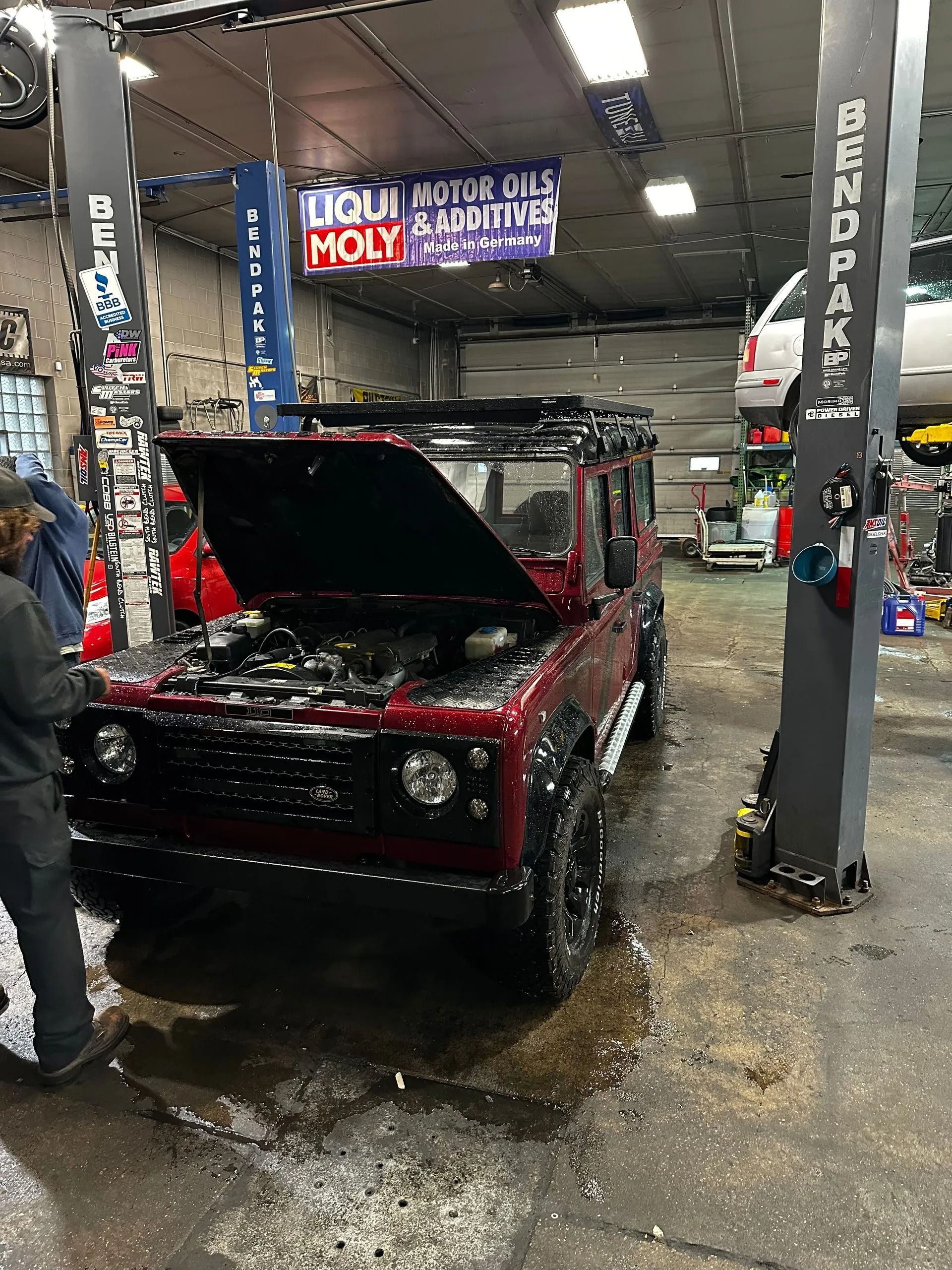 Red Land Rover with open hood in a garage on a lift; mechanic stands nearby.