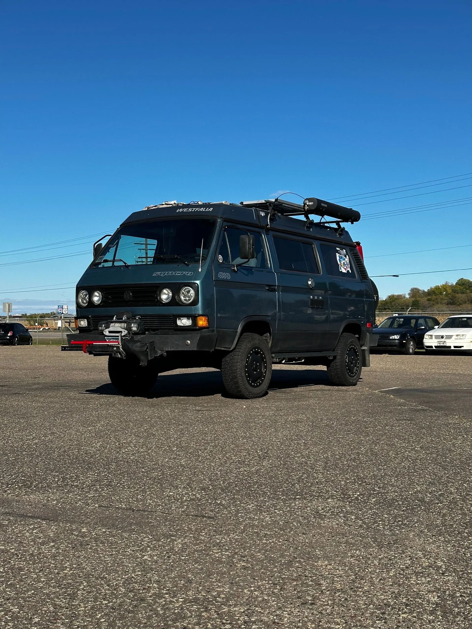 Dark blue lifted Volkswagen van with black wheels, roof rack, and bumper in a parking lot.