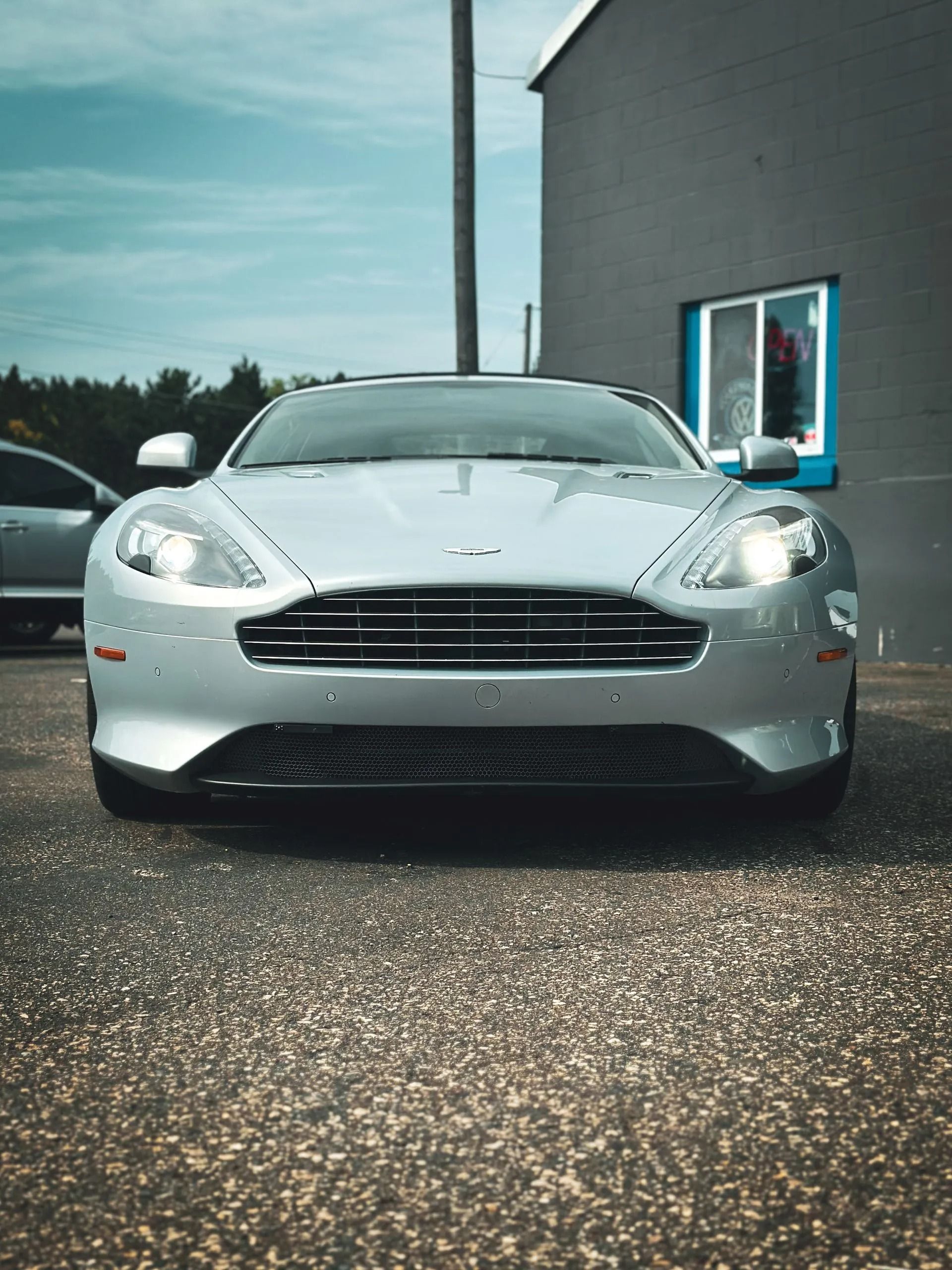 Silver Aston Martin sports car parked in front of a gray building on a sunny day.
