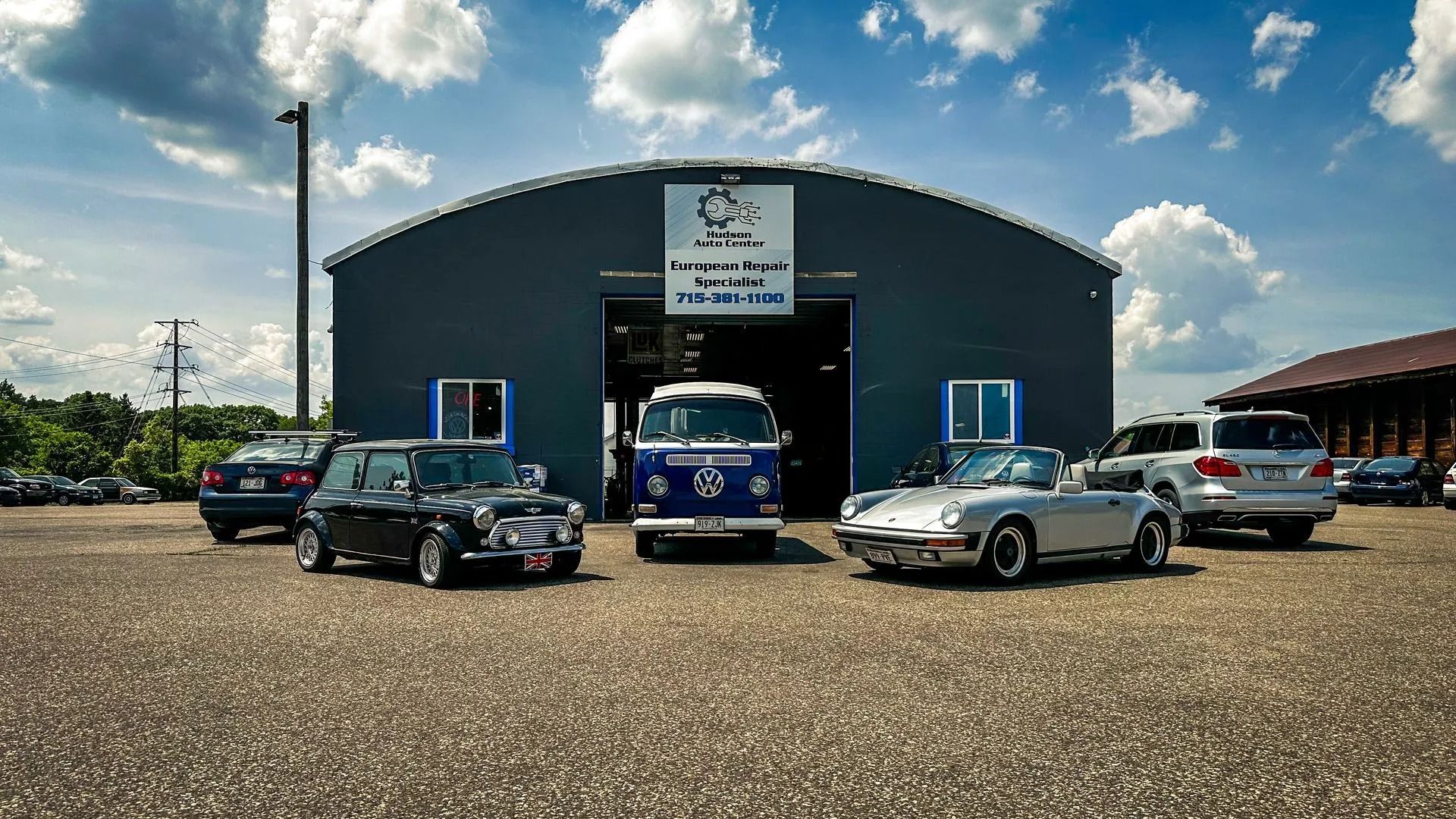 Cars parked in front of a garage with open door. A blue VW bus is centered, with a black Mini on the left and Porsche on the right.