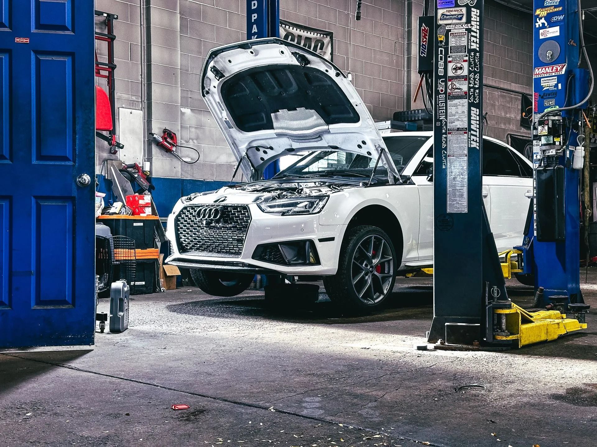 White car on a lift in a garage with hood open, blue door on the left, gray floor.
