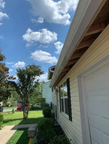 Side view of a house with a white garage door, light siding, and a gutter against a blue sky with clouds.
