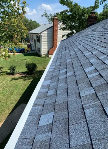 View of a blue shingled roof with a white gutter, and a house in the background.