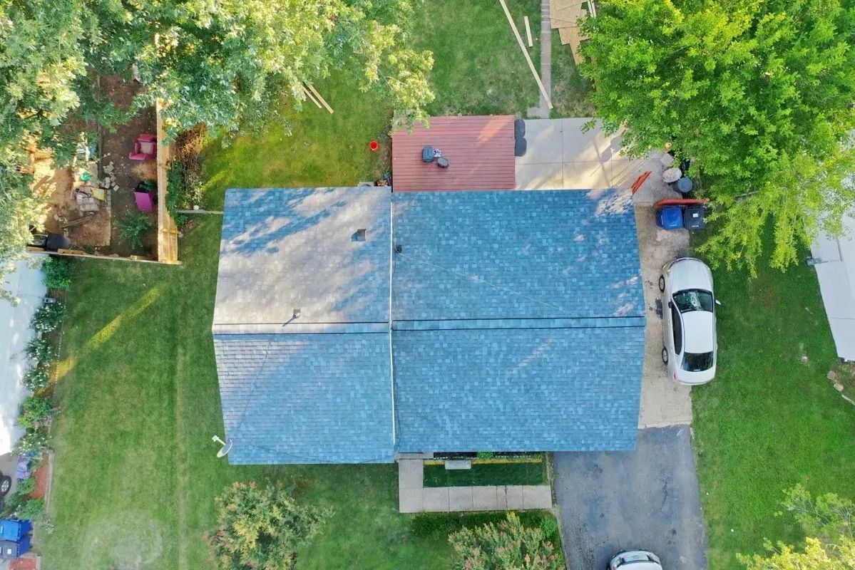 Overhead view of a blue-roofed house with a deck, driveway, and parked white car. Green lawn surrounds.