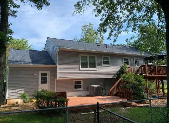 Back of a two-story house with gray siding, dark roof, wooden deck, and a fenced yard under a sunny sky.