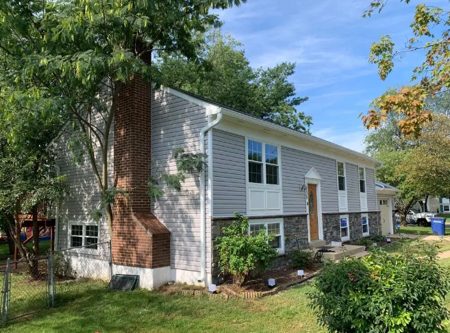 Two-story house with gray siding, brick chimney, stone accent, and front door, trees and blue sky.