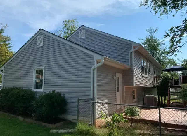Gray-sided house with a white window and door, surrounded by greenery and a black fence, under a blue sky.