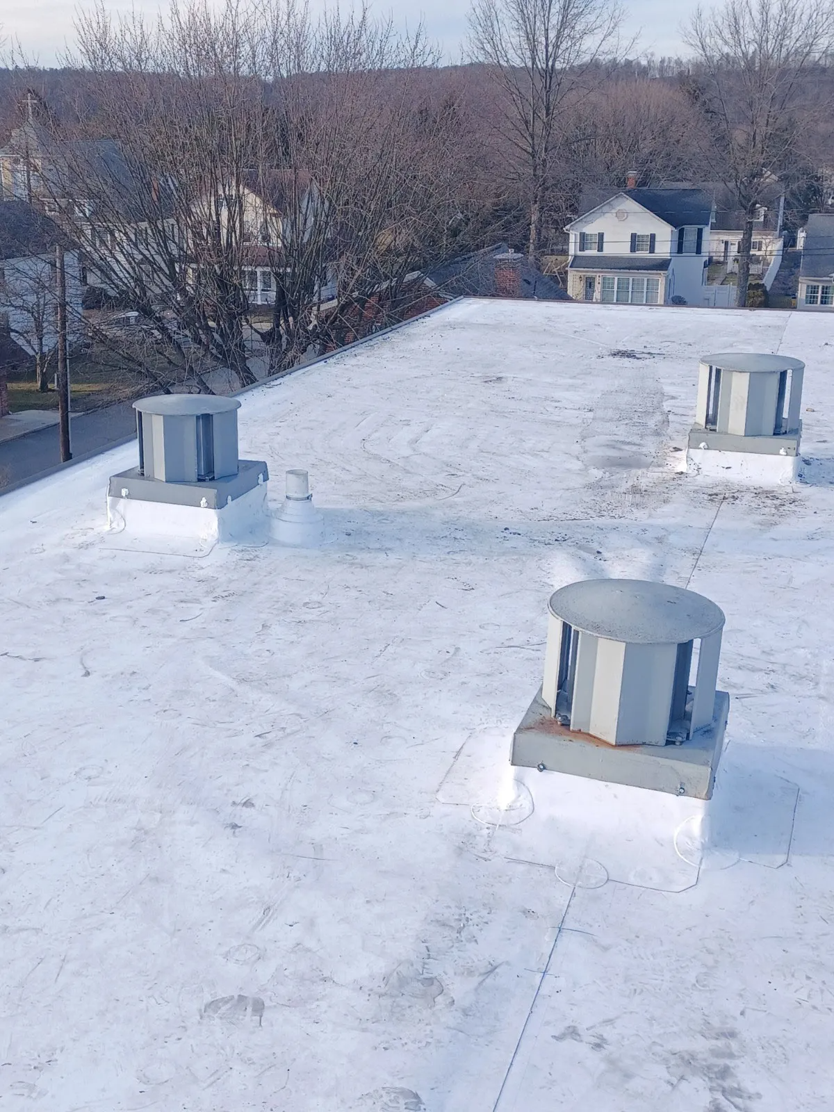 Snow-covered rooftop with three metal vents and trees and houses in the background