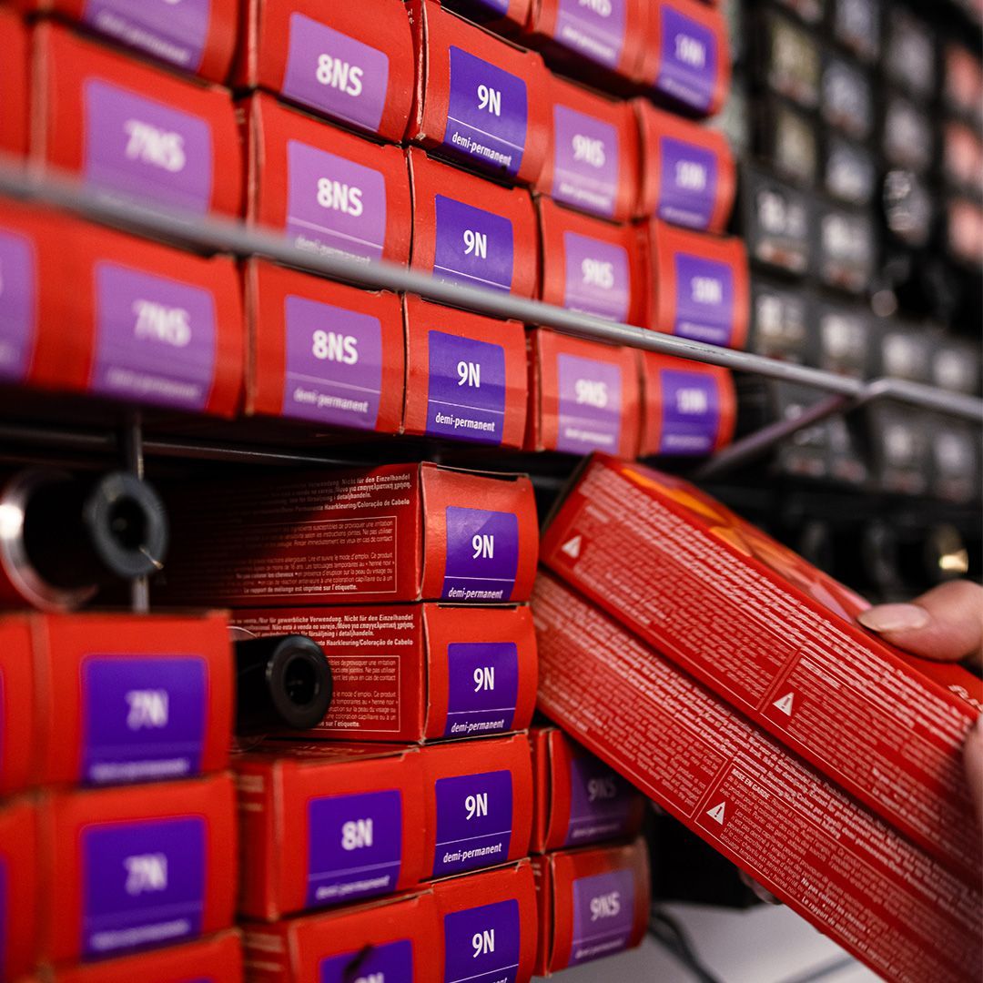 A hand selects a red box of hair dye from a shelf filled with organized, labeled boxes of professional hair color products.