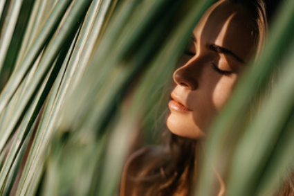 A person with their eyes closed, partially obscured by blurred, sunlit palm fronds, in a close-up portrait.