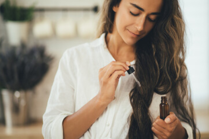 A person in a white shirt holding a small amber glass bottle and dropper, about to apply serum to their long, wavy hair.