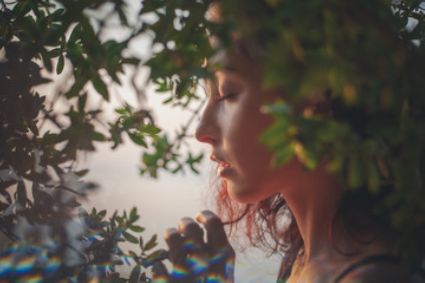 Profile view of a person with eyes closed, surrounded by foliage with soft, warm lighting and a colorful light flare.