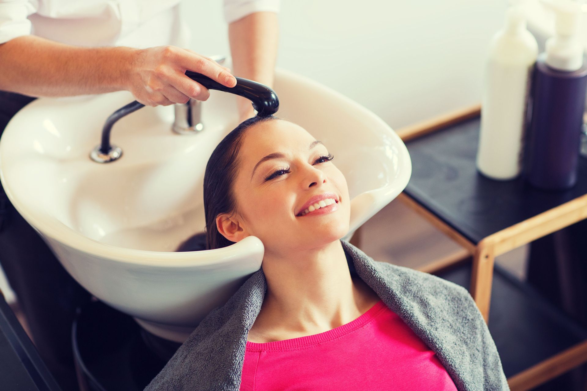 A person with a smiling expression gets their hair washed in a salon sink by a professional.
