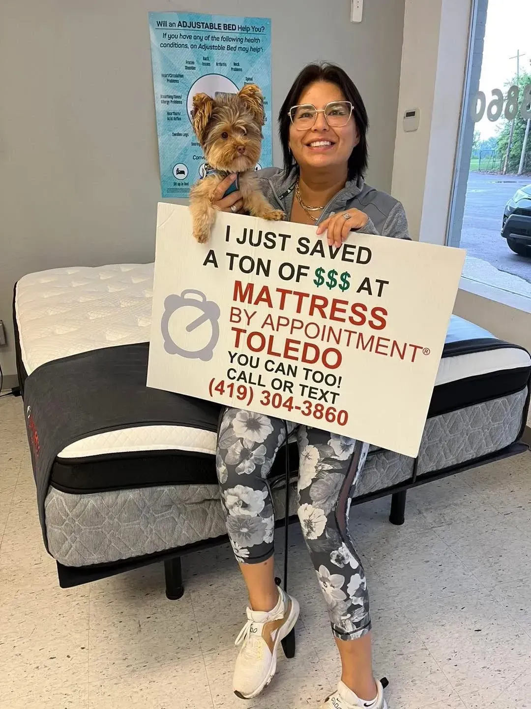 Woman with dog holding sign in front of a mattress. Sign says, 
