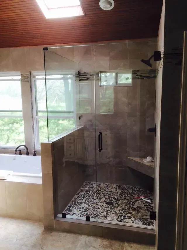 Glass-enclosed shower with pebbled floor, built-in seat, dark fixtures, and light-colored tile in a bathroom with skylight.