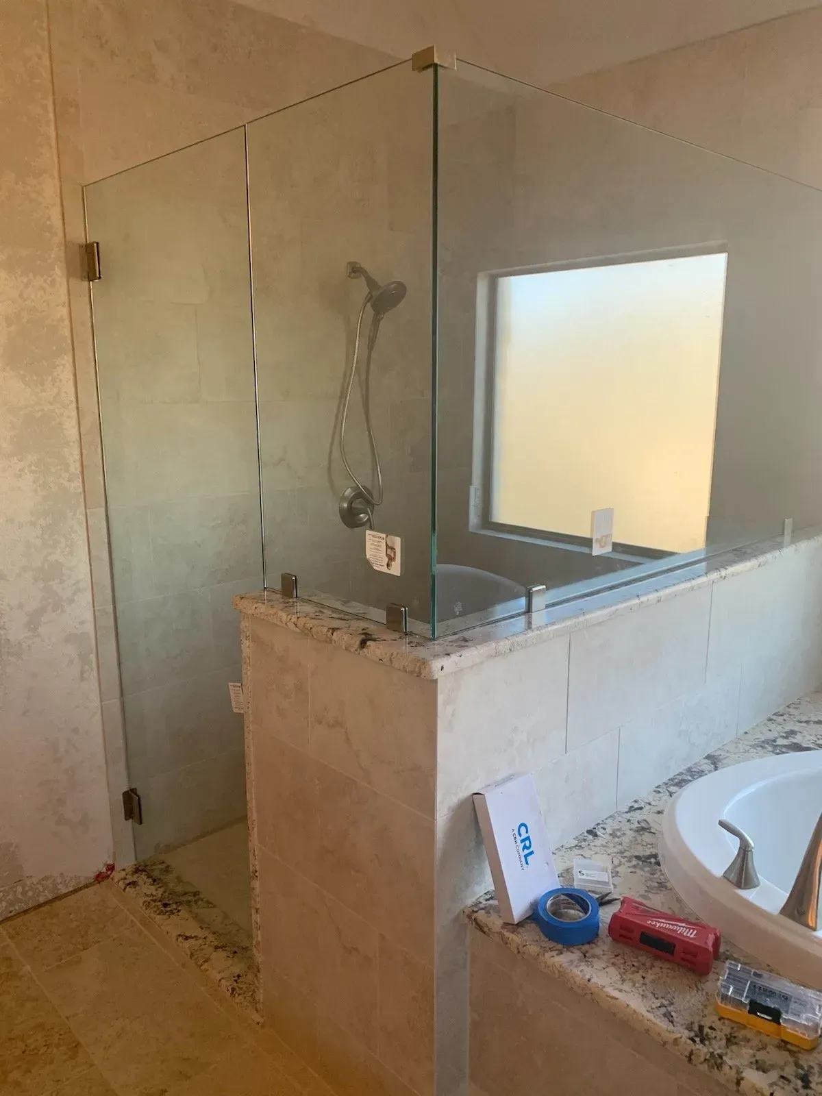 Glass shower enclosure in a bathroom with a window and a granite countertop.