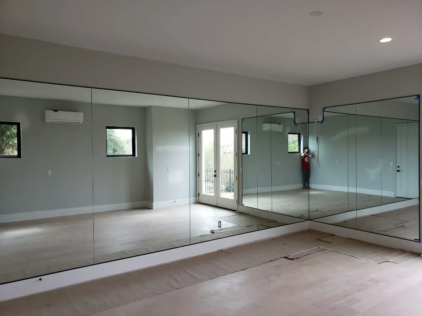 A room with mirrored walls, light wood floor, and two windows. A person is visible in the mirror.