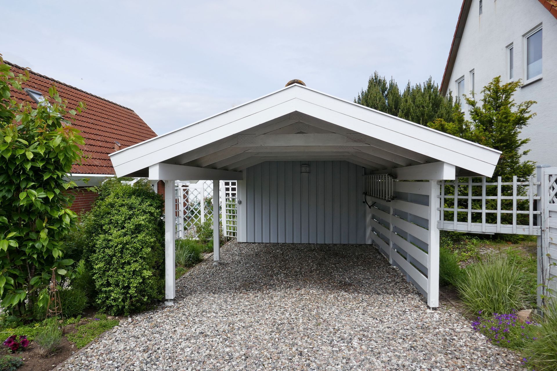 White carport with gravel driveway, light blue siding, and white fence in front of a house.