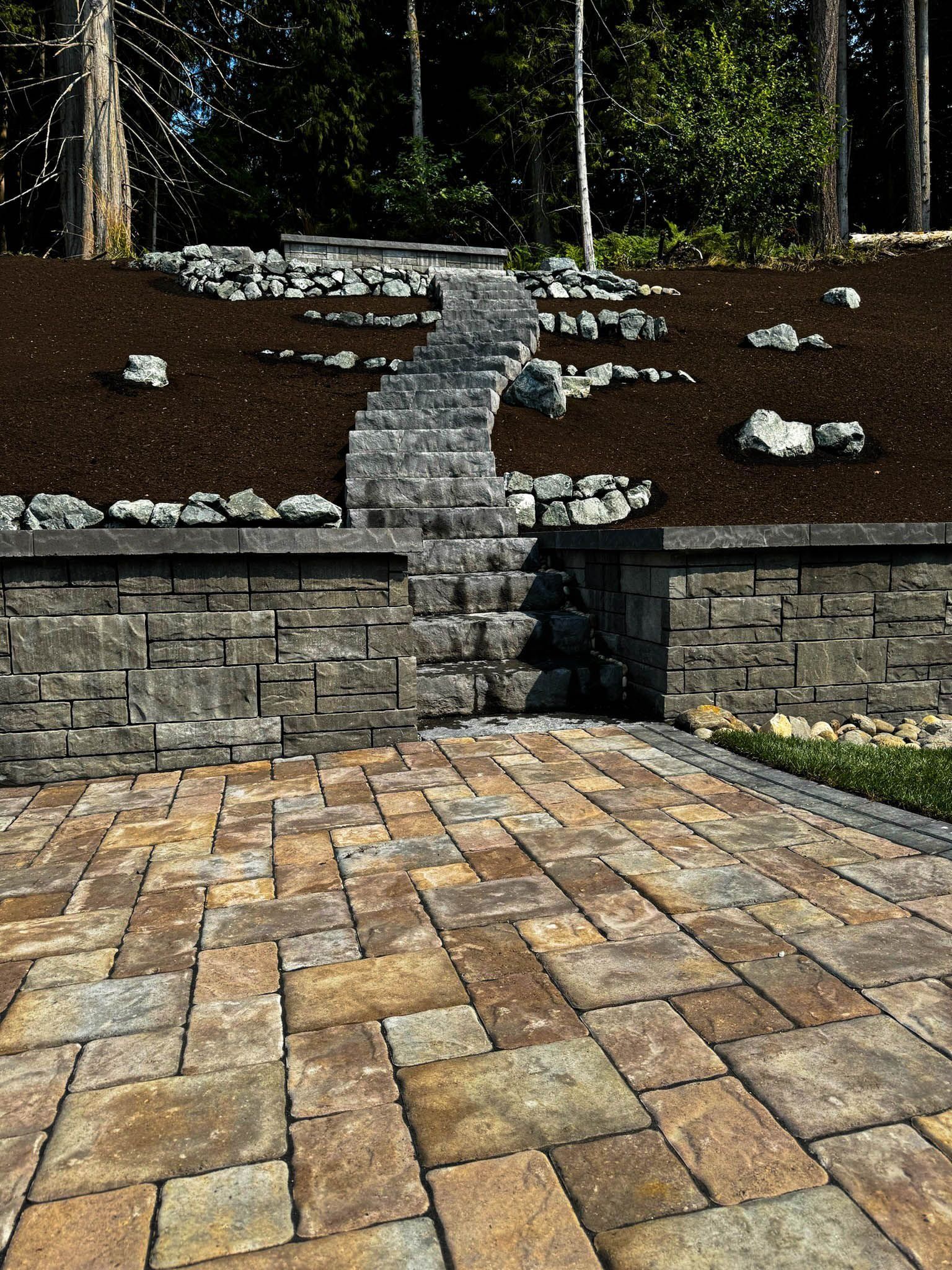 Stone patio with steps leading up a slope. Retaining walls, mulch, and trees in background.