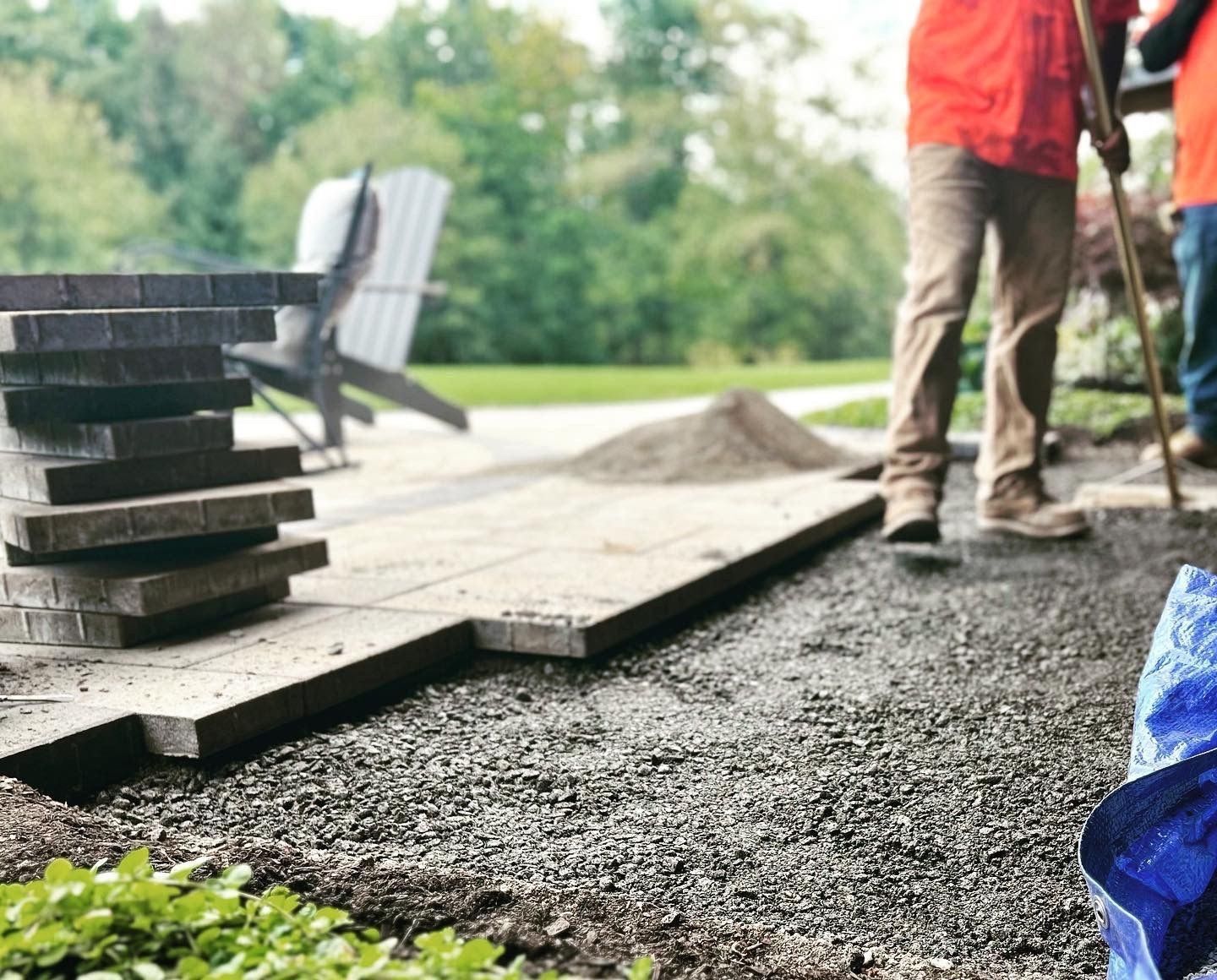 Workers laying patio pavers outdoors; gravel base, stacked pavers, red shirt, sunny day.