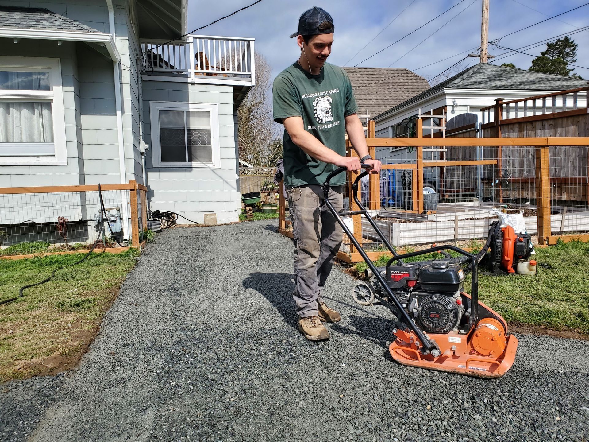 Man using a plate compactor on a gravel driveway next to a house with a blue facade.