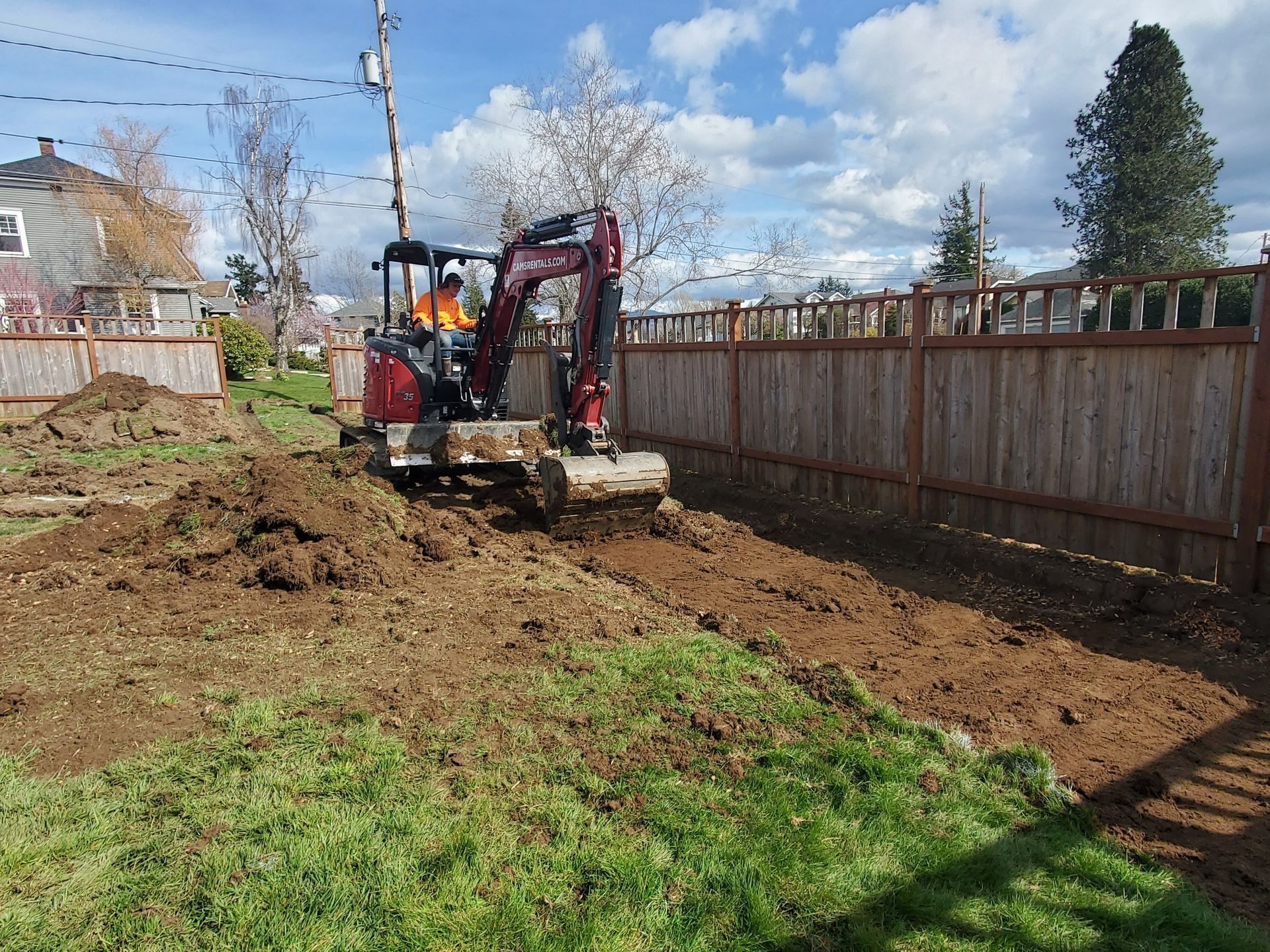 Mini excavator digging in a grassy yard next to a wooden fence.