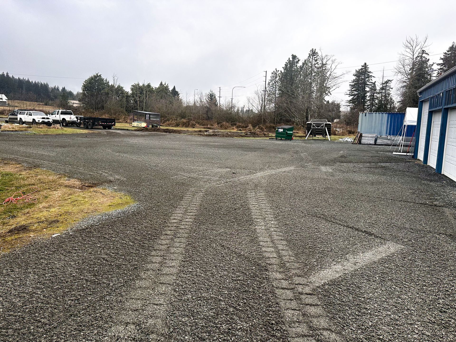 Gravel lot with tire tracks, parked vehicles in background, and storage units to the right under a cloudy sky.