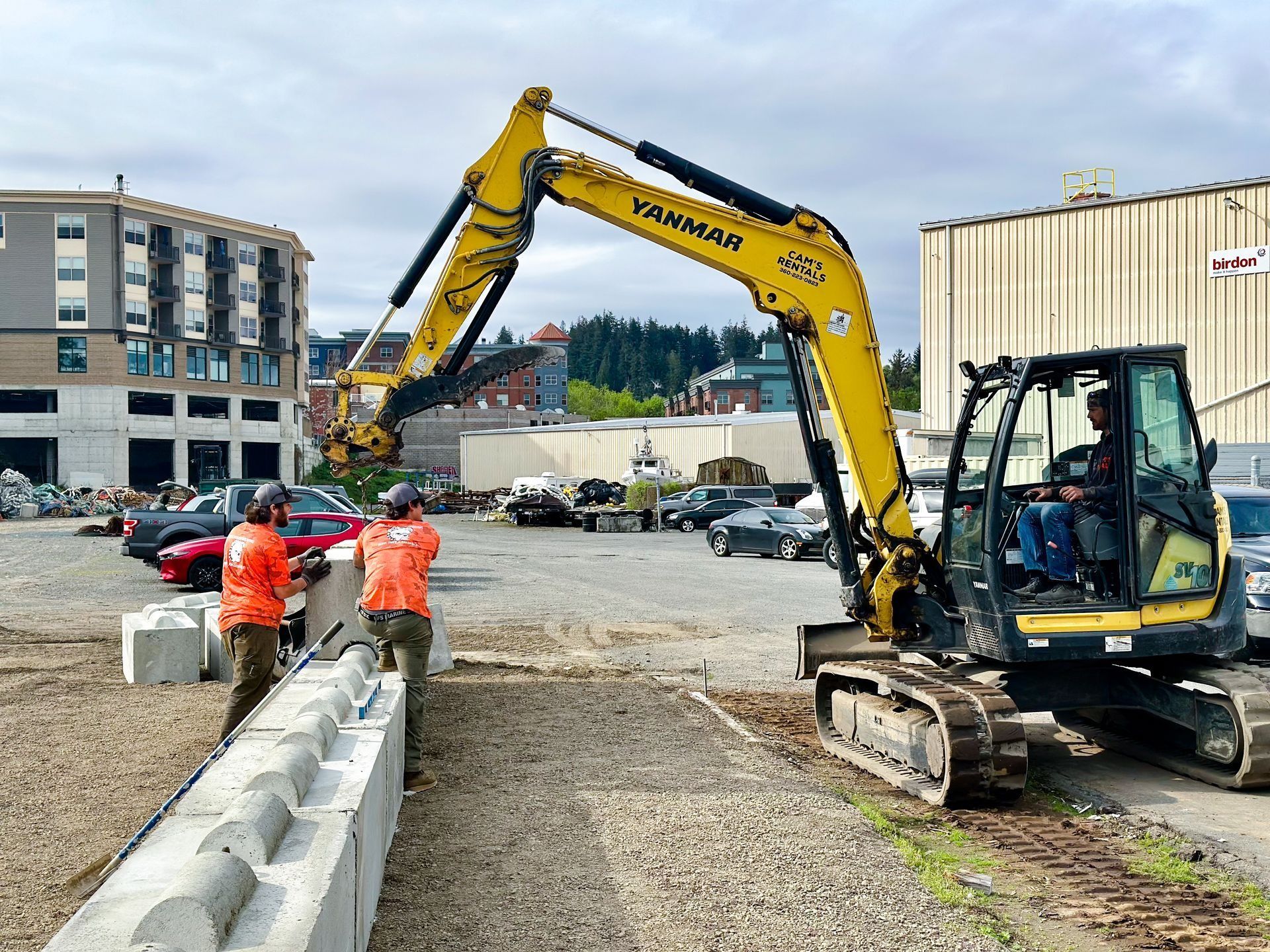 Yellow excavator placing concrete blocks; two workers in orange vests watch.