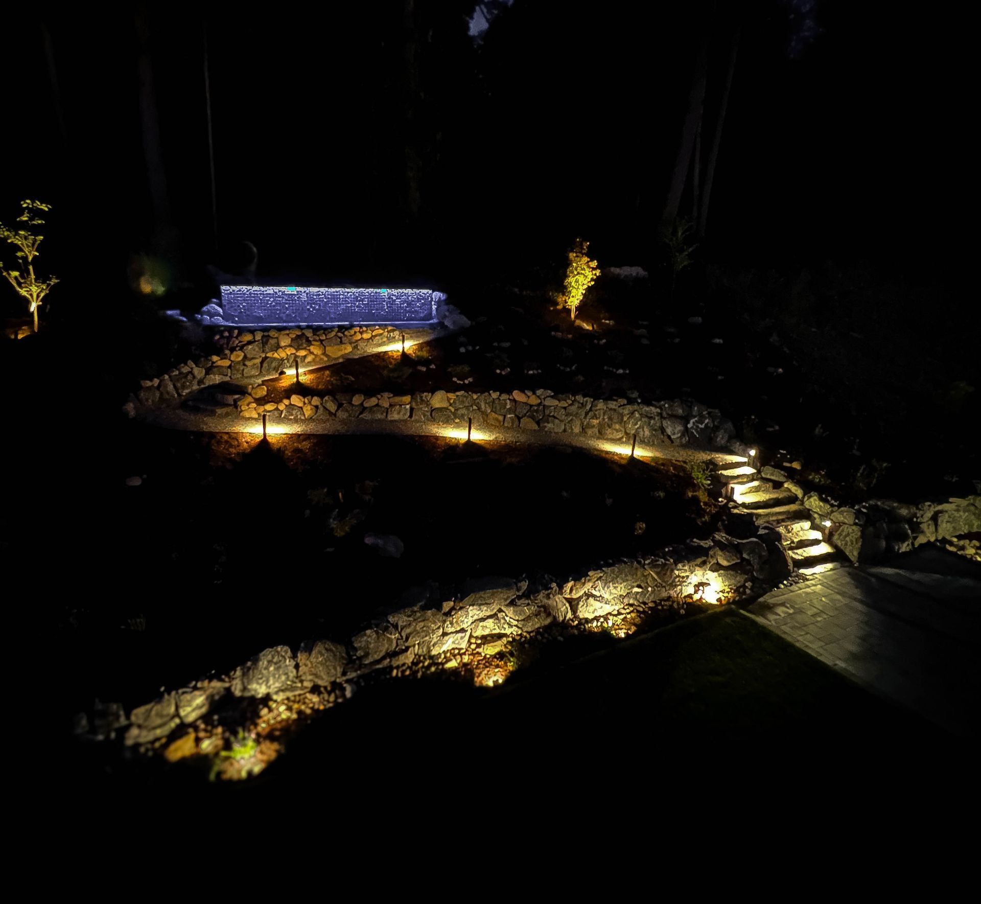 Nighttime garden path with landscape lighting illuminating stone walls, steps, and greenery.
