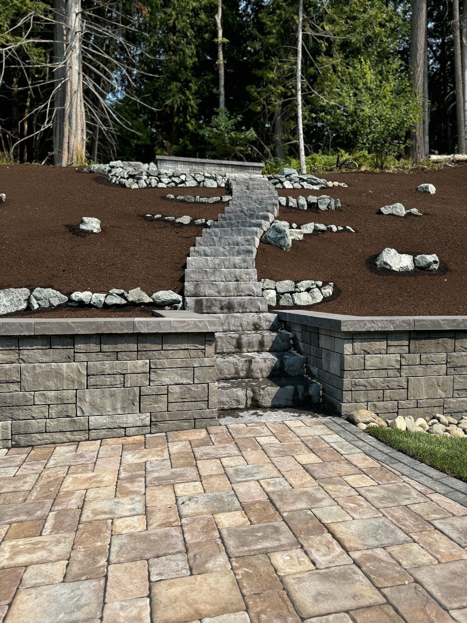 Stone steps leading up a hillside, framed by a retaining wall and a paved patio. Trees in the background.