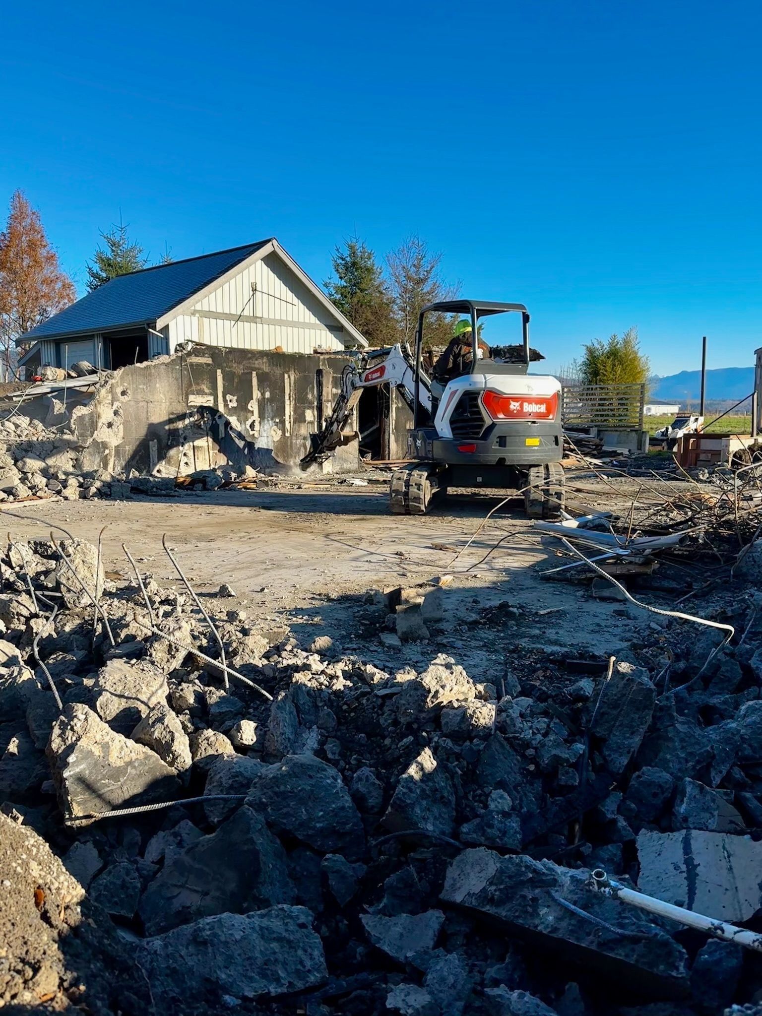 Bobcat excavator demolishing a building; rubble in foreground, blue sky overhead.
